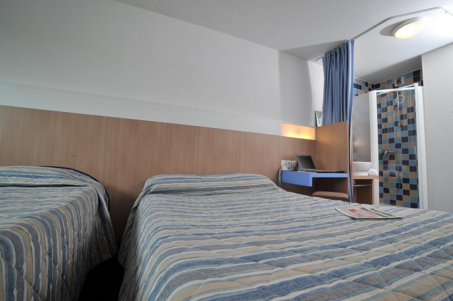 Ein Hotelzimmer mit zwei Betten, einem Schreibtisch mit einem Laptop, einer Dusche und einem Spiegel.