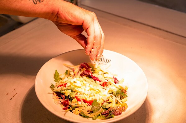 Una mano esparciendo condimentos sobre una ensalada en un tazón blanco.