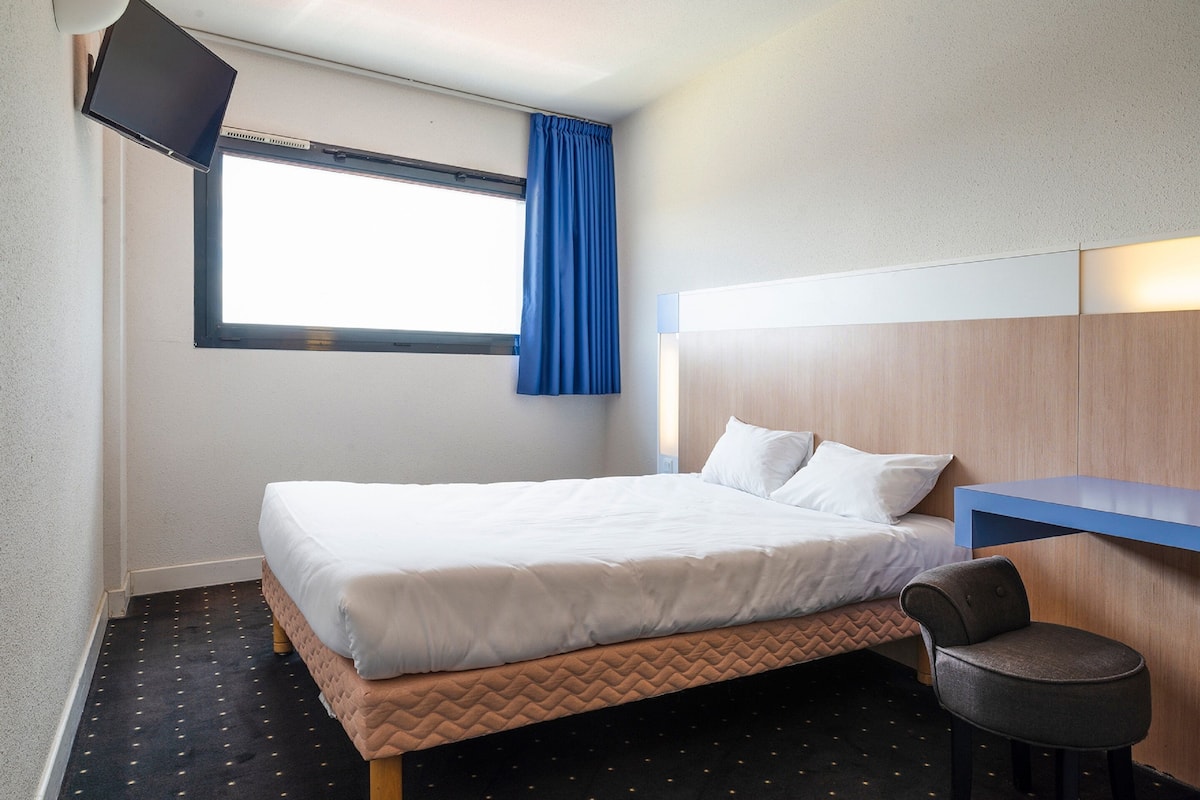 Ein Hotelzimmer mit Bett, Schreibtisch, Stuhl, Fernseher und einem Fenster mit Vorhängen.