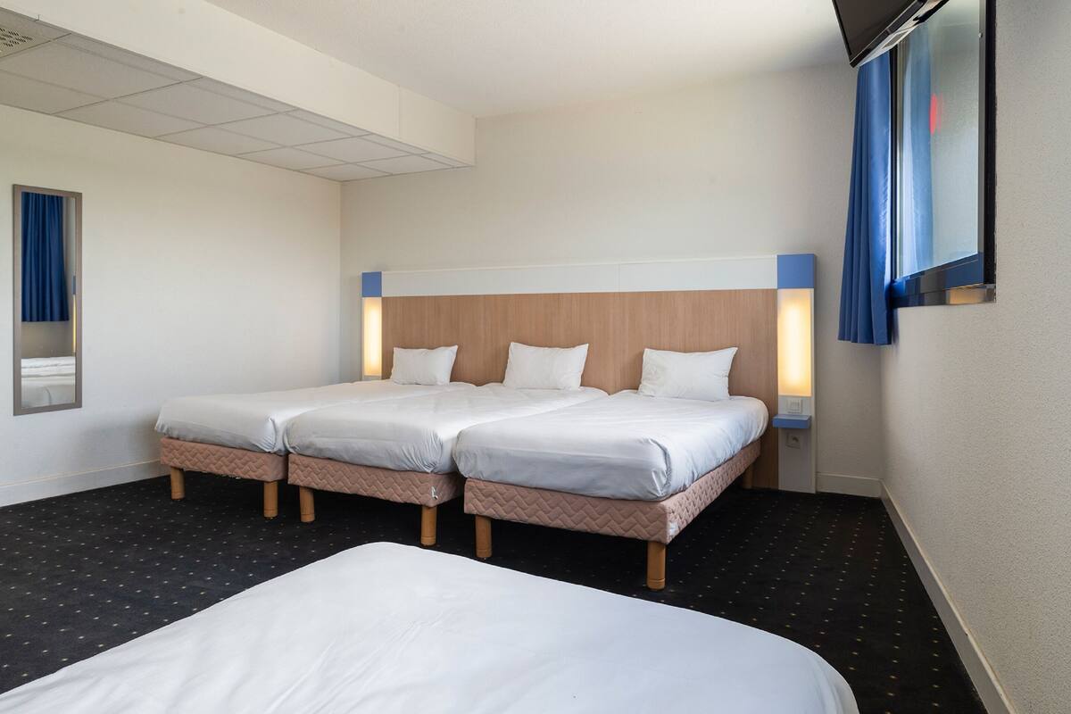 Ein Hotelzimmer mit zwei Betten, ein großes Bett im Vordergrund und ein kleineres im Hintergrund. Ein Fernseher ist an der Wand befestigt und es gibt ein Fenster mit Vorhang.