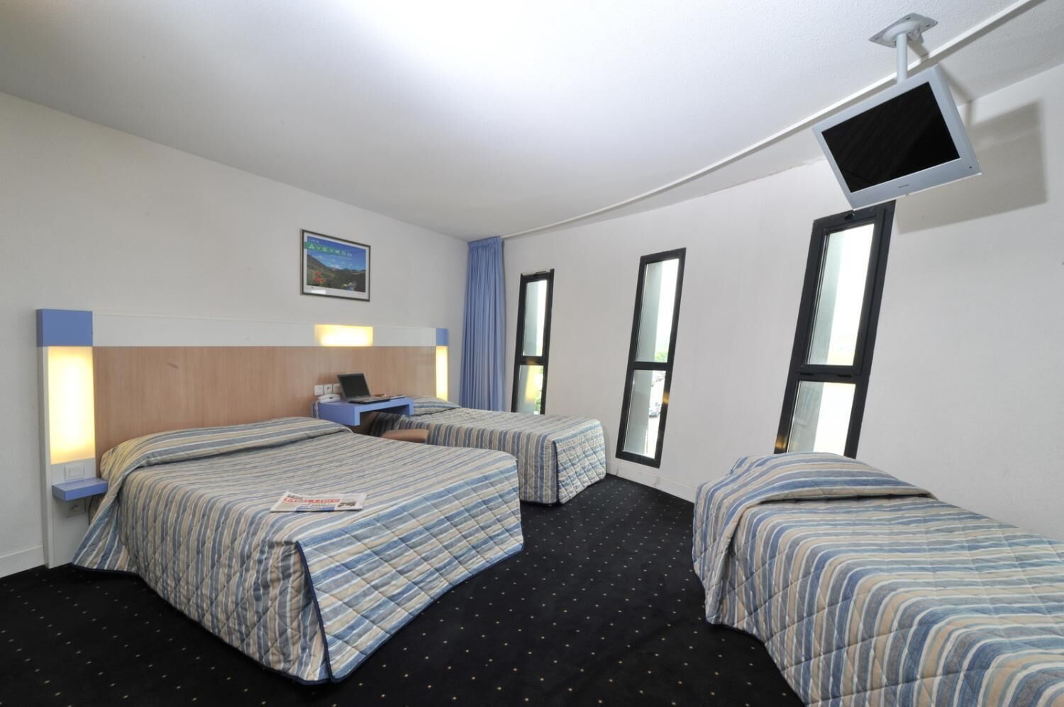 A hotel room with two beds, a desk with a laptop, a flat-screen TV mounted on the wall, and a window with curtains.