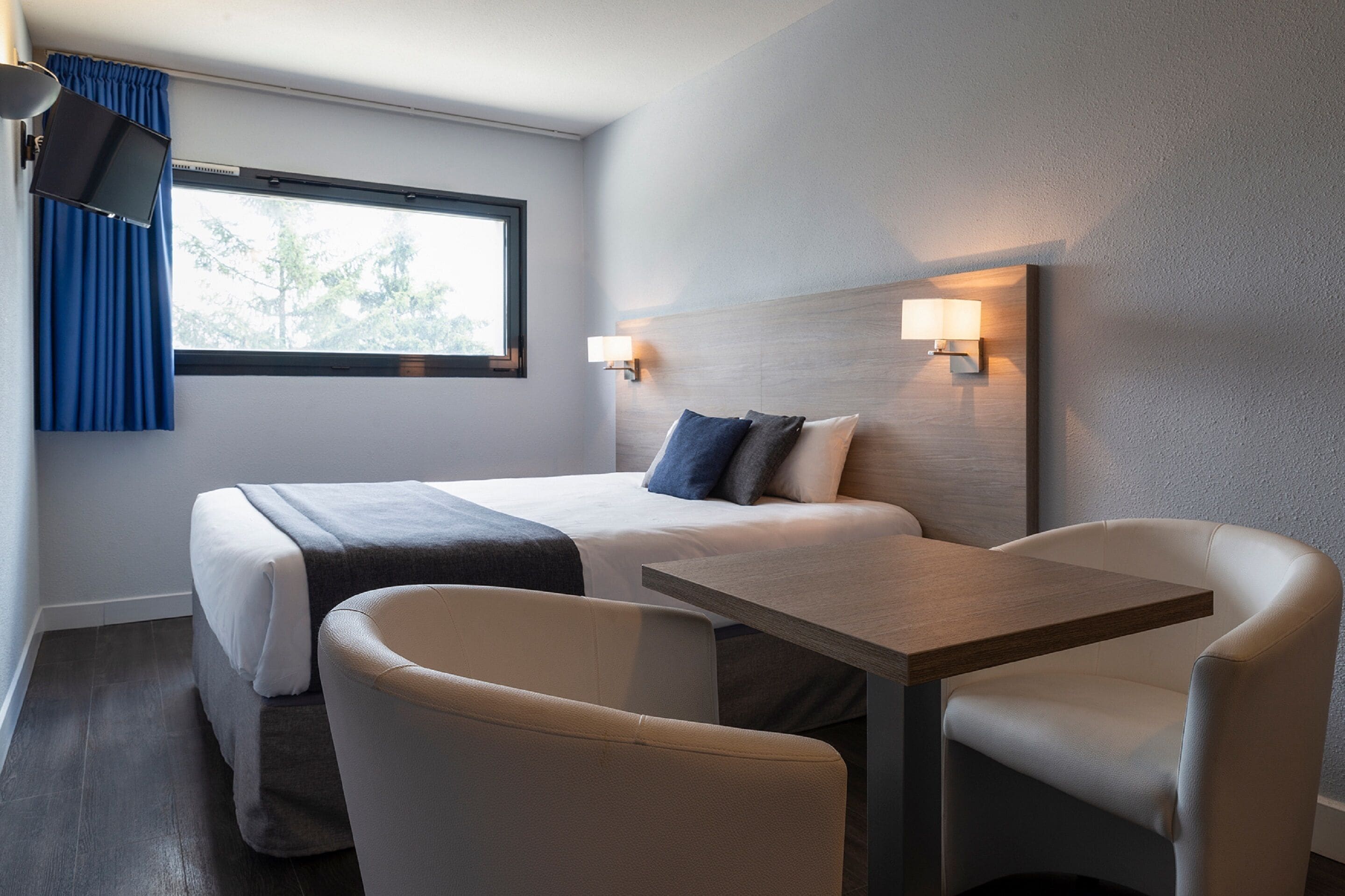 Een hotelkamer met een bed, twee stoelen, een tafel, een televisie en een raam met gordijnen.