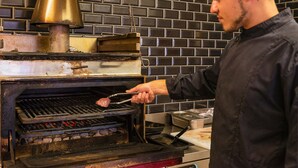 A chef cooking meat in a brick-walled kitchen with a large brass stove.