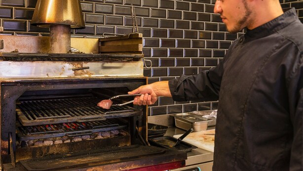 Ein Koch bereitet Fleisch in einer Küche mit Backsteinwänden und einem großen Messingherd zu.