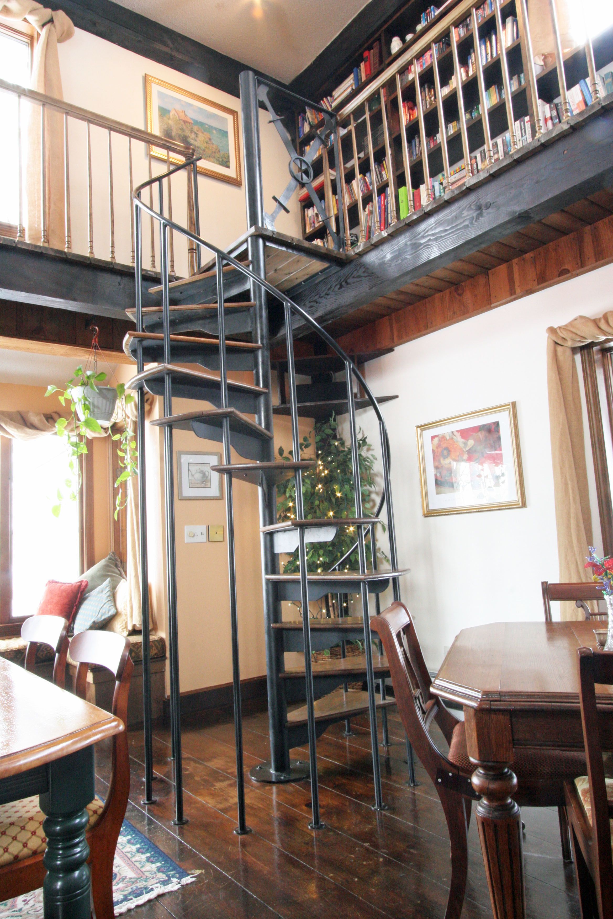 A dining area with a wooden table and chairs, a spiral staircase, and a bookshelf with various items.