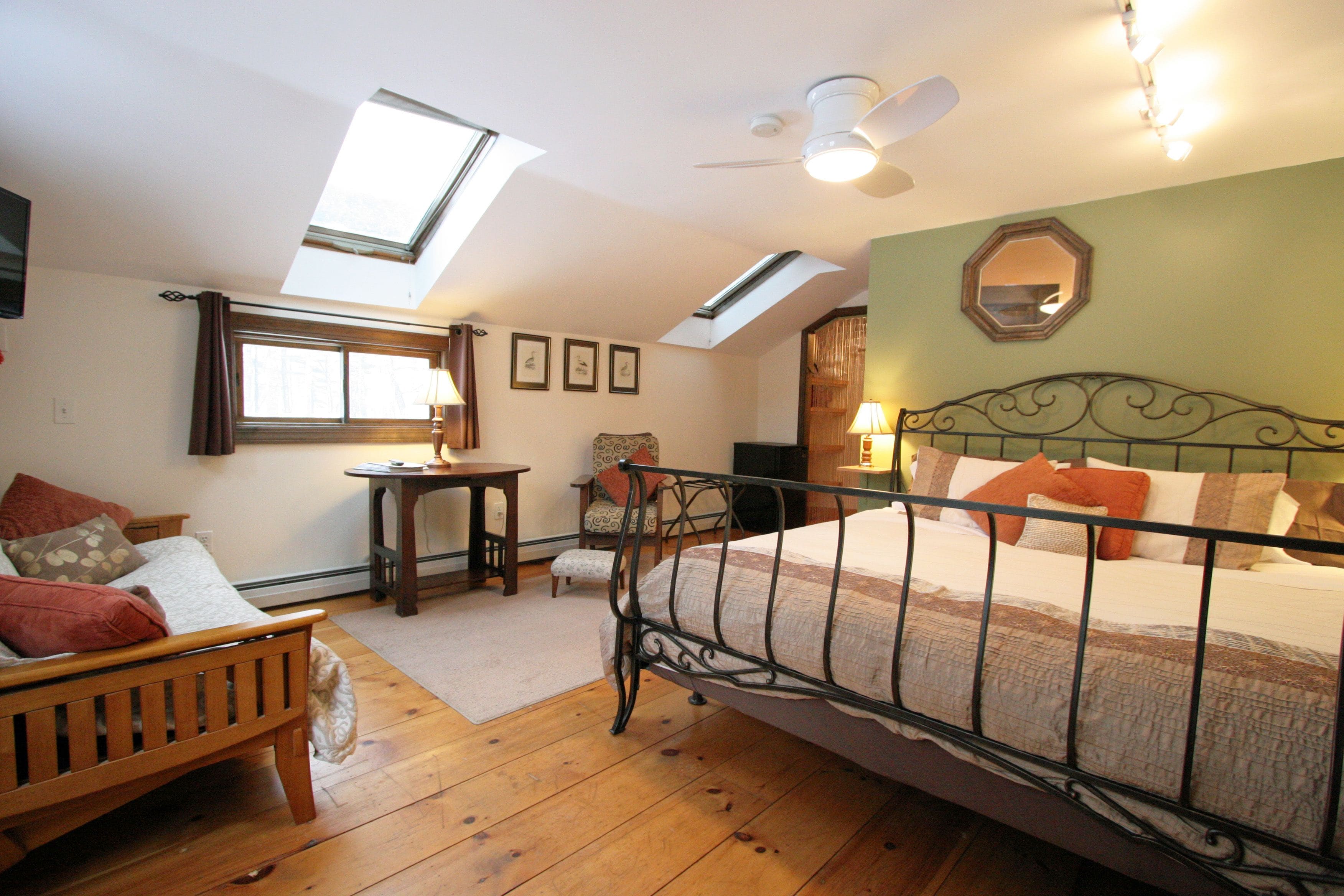 A bedroom with a sloped ceiling, skylight, a bed with a metal frame, a wooden bench, a small table, and a chair.