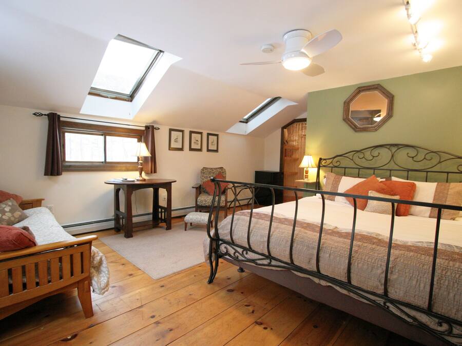 A bedroom with a sloped ceiling, skylight, a bed with a metal frame, a wooden bench, a small table, and a chair.