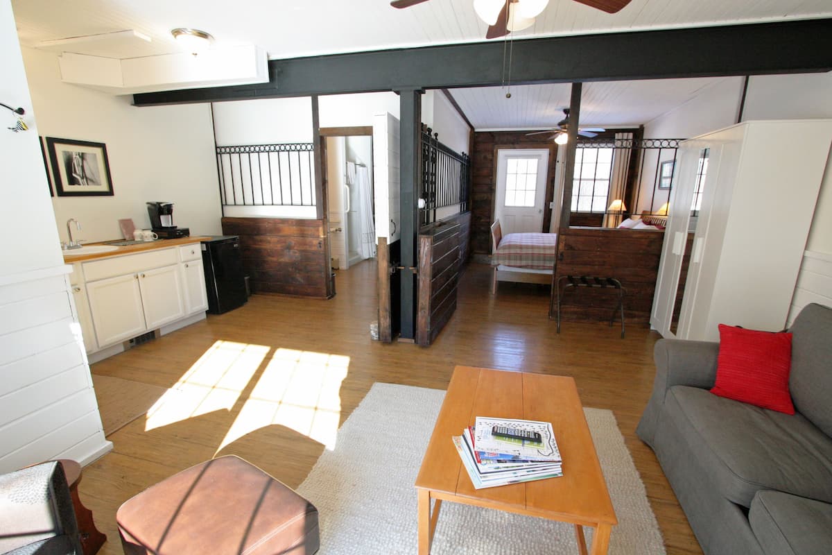 A spacious living area with a sofa, a coffee table, and a dining table. There is a kitchen area with cabinets and a refrigerator. The room has wooden flooring and a ceiling fan.
