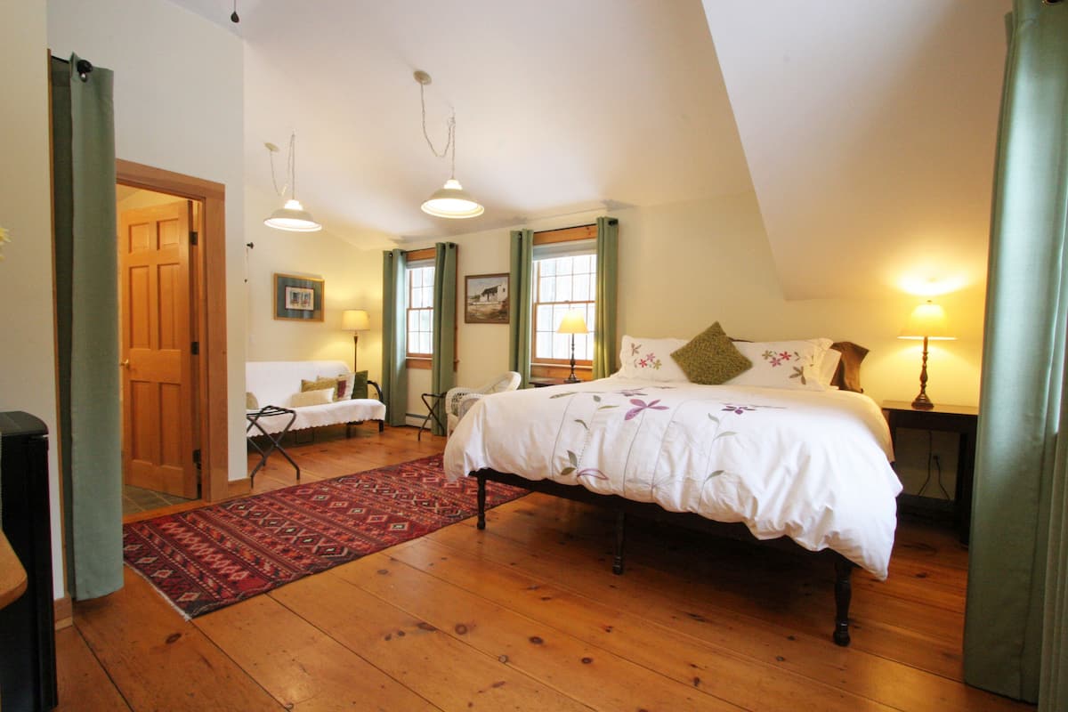 A bedroom with a wooden floor, a bed with a white bedspread, two windows with green curtains, a red rug, and a wooden door.