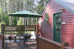 A wooden deck with a table and chairs, a green umbrella, and a red building with a green door.
