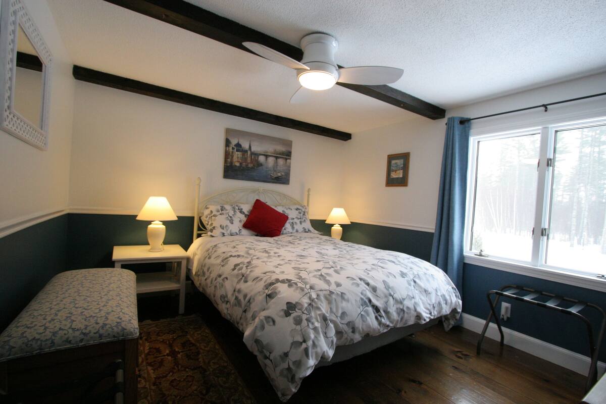 A bedroom with a bed, two bedside tables with lamps, a bench, a ceiling fan, and a window with curtains.