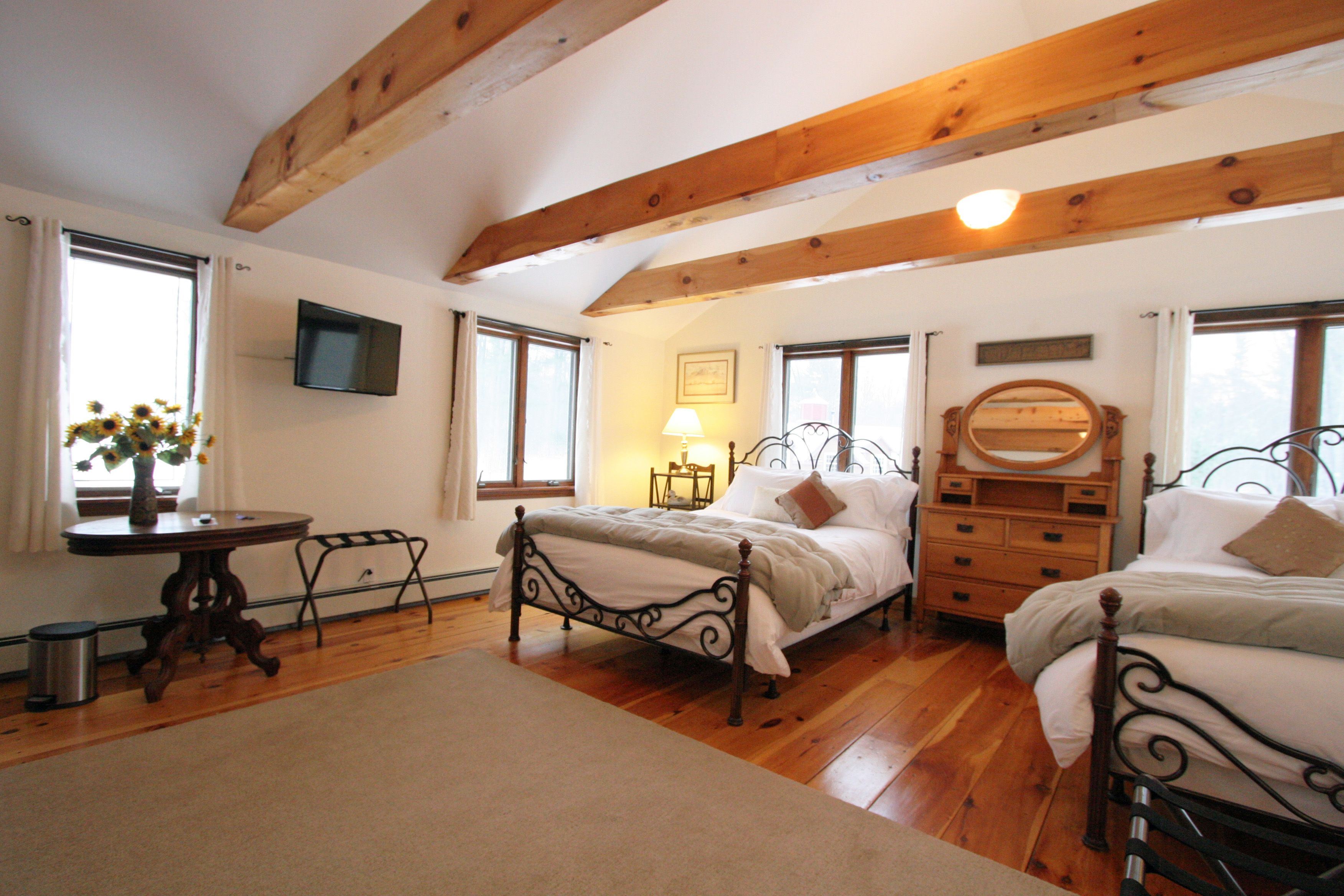 A bedroom with two beds, a wooden ceiling, a TV, a table with a vase of flowers, and a lamp.