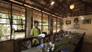A dining area with a long table set for a meal, large windows with views of greenery, and a person standing by the table. - Grassroots Wayanad, Valley-view Tents (Vythiri)
