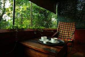 A wooden bench with a tray holding two cups and a small container, set against a window overlooking lush greenery. - Grassroots Wayanad, Valley-view Tents (Vythiri)