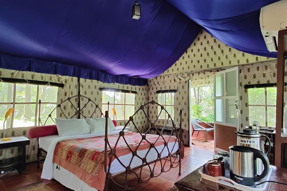 A tented room with a bed, a table with a kettle, and a window with a view of greenery.