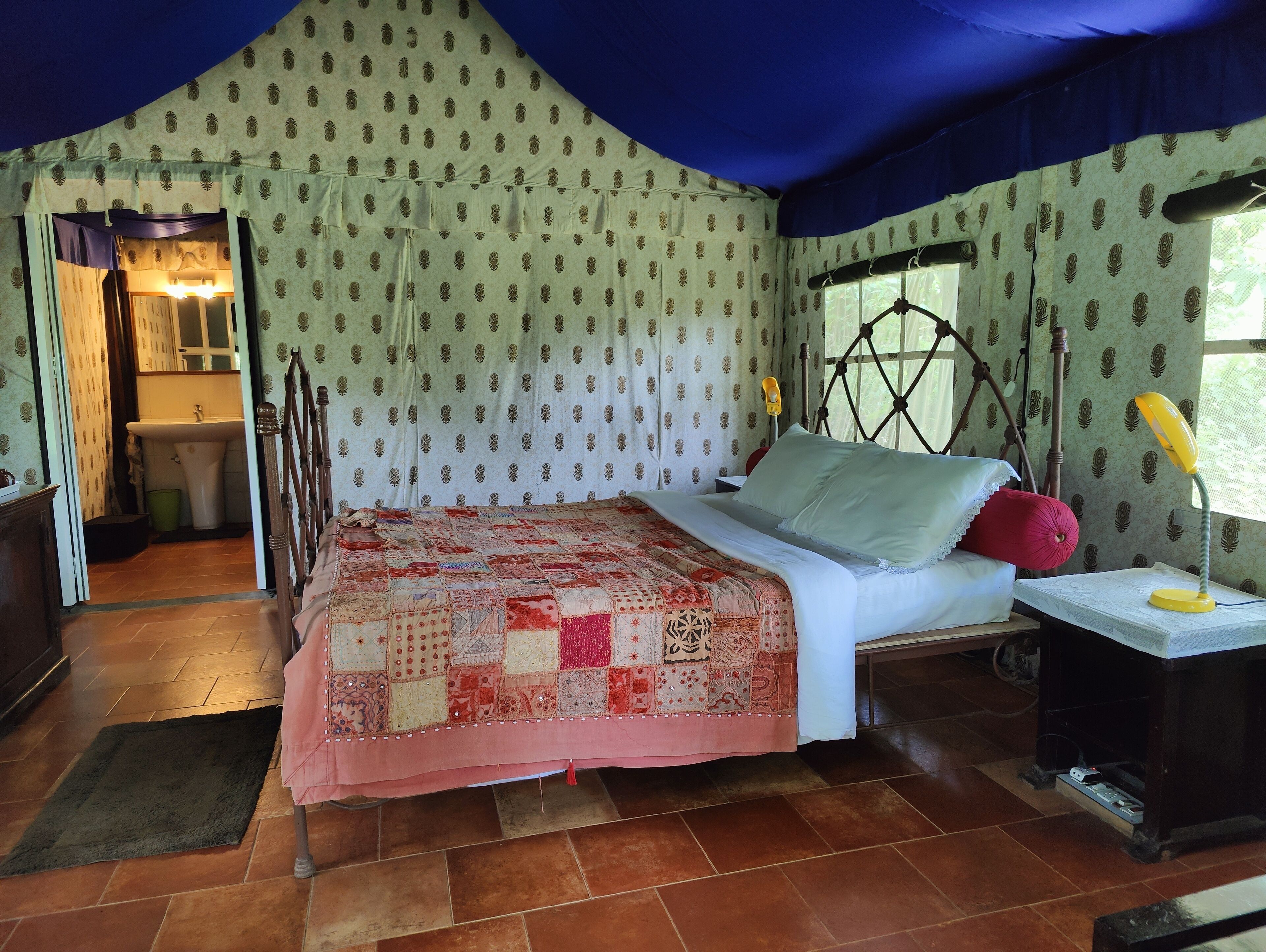 A bedroom with a bed, a nightstand, a window with curtains, a wooden headboard, and a bathroom visible through an open door.