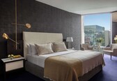 A modern hotel room with a large bed, a nightstand, a lamp, a chair, and a view of the city through large windows.
