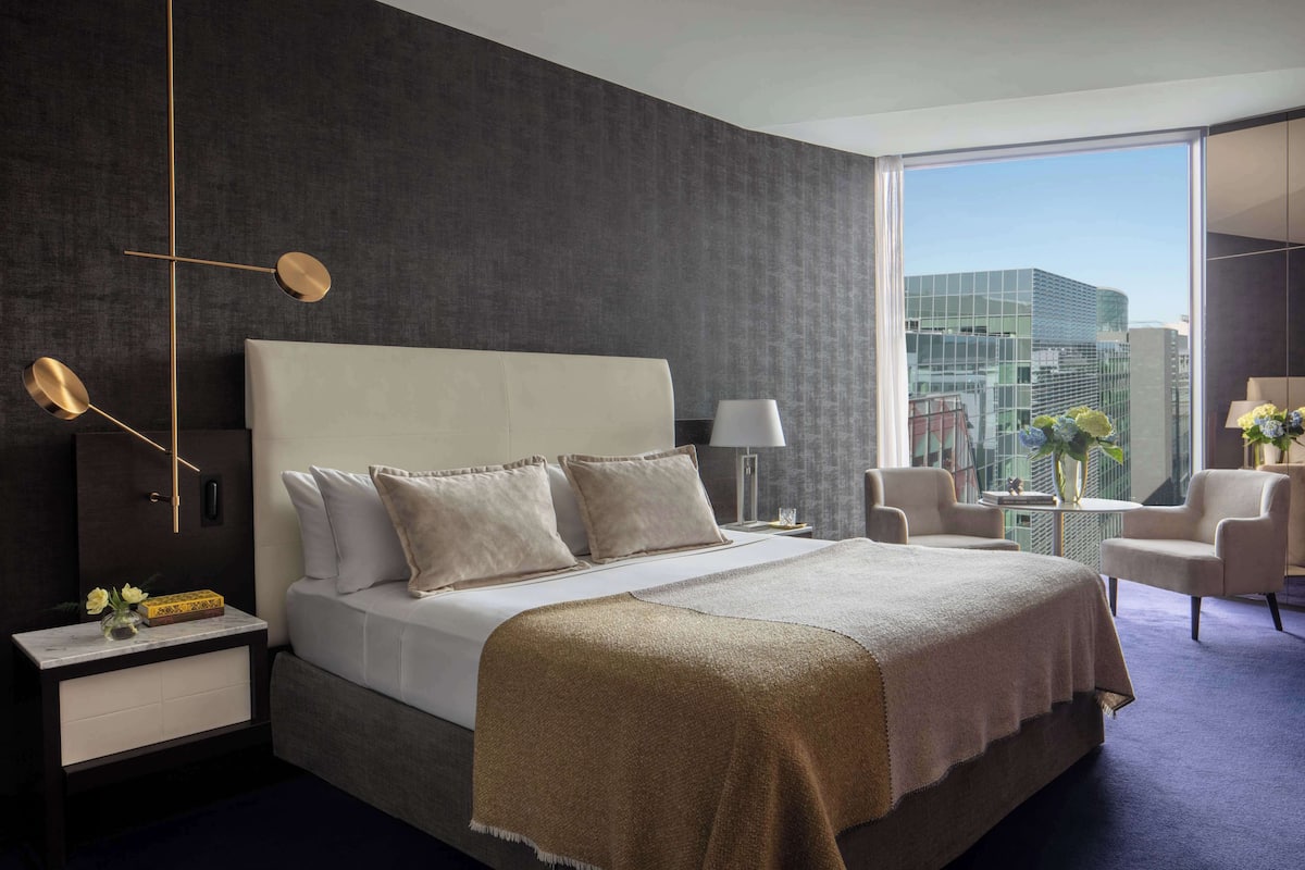 Una habitación de hotel moderna con una cama grande, una mesita de noche, una lámpara, una silla y vistas a la ciudad a través de amplios ventanales.