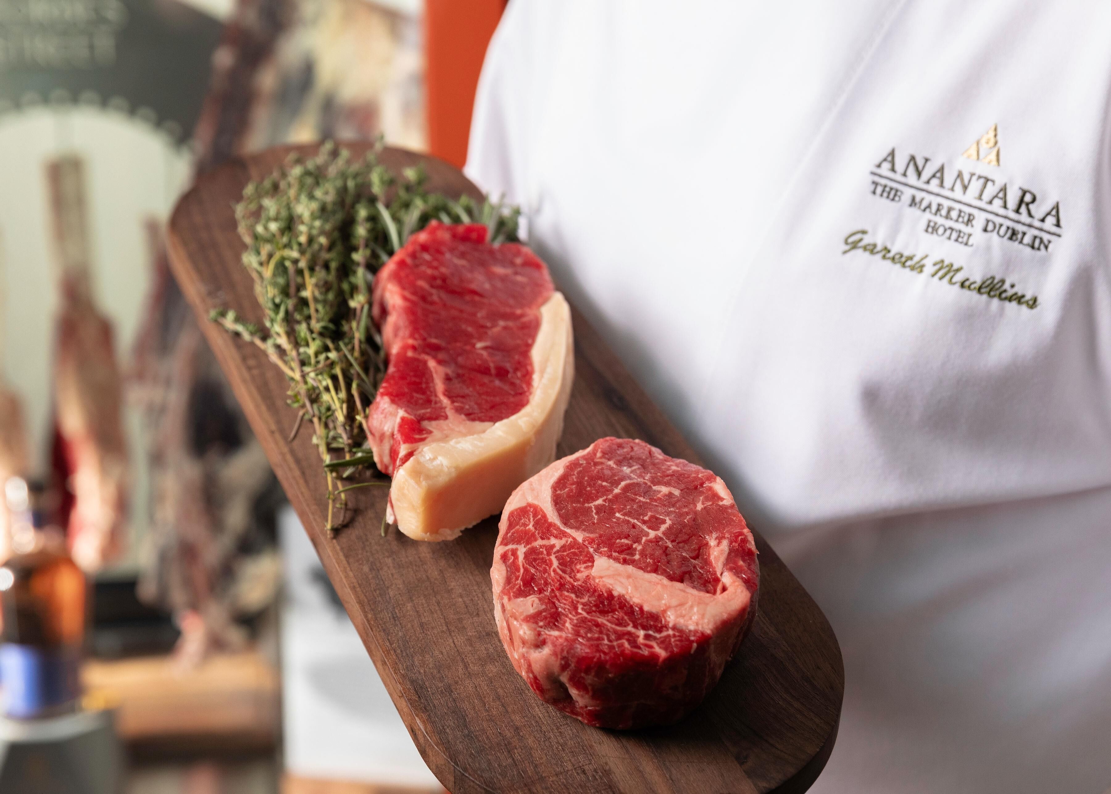 A person in a white apron holding a wooden board with two raw steaks and some herbs. The text on the apron reads "ANANTARA THE MARKER DUBLIN GARTH MULLINS."