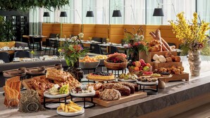 Una mesa de buffet con panes, quesos, frutas y pasteles en un moderno comedor con ventanas amplias y plantas de interior.