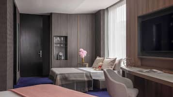 A modern hotel room with a large bed, a desk with a chair, a TV, and a small seating area with a sofa and a table.