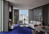 A modern living room with a sofa, armchairs, a coffee table, and a dining table with fruit. There is a large abstract painting on the wall and a view of buildings through the window.
