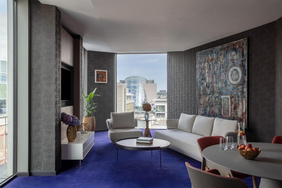 Una sala moderna con sofá, sillones, una mesa de centro y una mesa de comedor con fruta. Hay un cuadro abstracto grande en la pared y vistas de edificios a través de la ventana.
