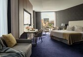 A hotel room with a bed, a sofa, a desk, and a view of the cityscape.