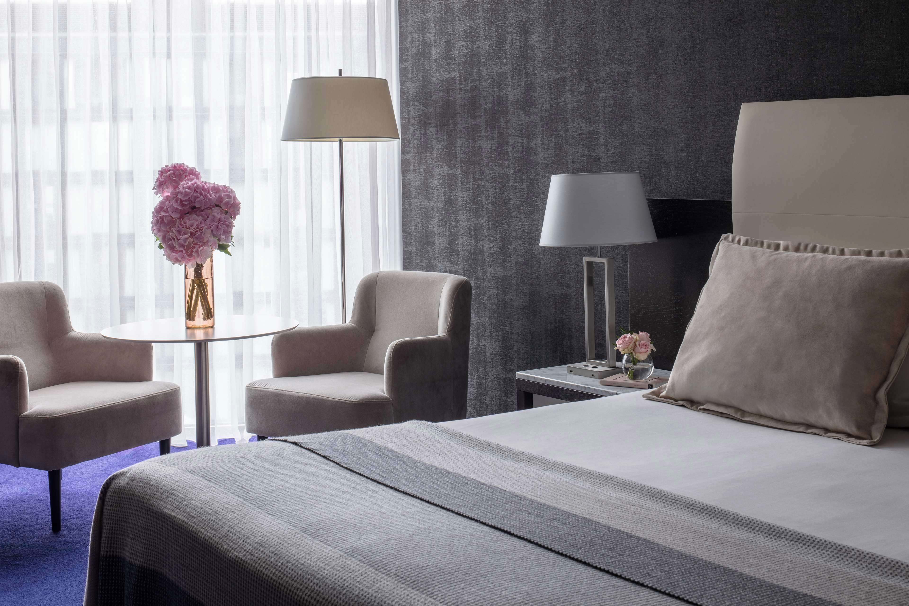 A hotel room with a bed, two armchairs, a small table with a vase of flowers, and a lamp.