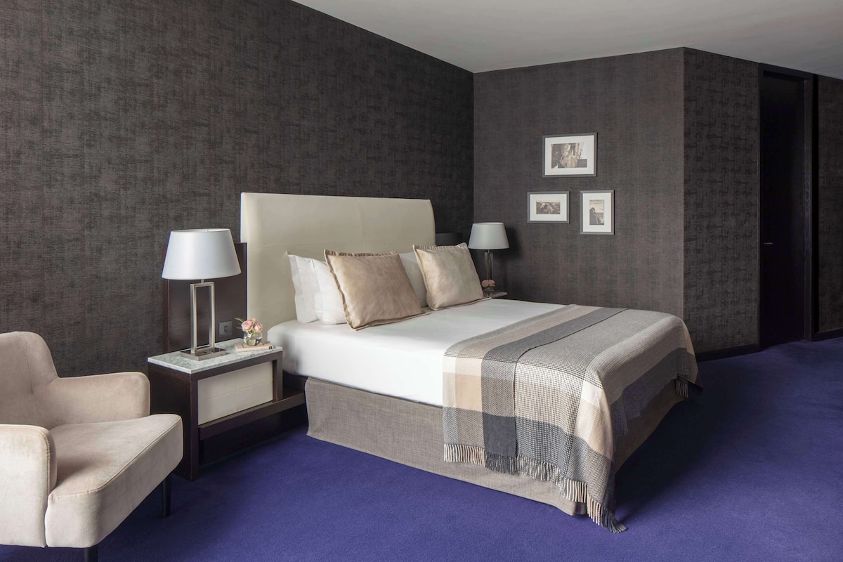 Una habitación de hotel moderna con una cama grande, dos mesitas de noche, una silla y una pared oscura con textura.
