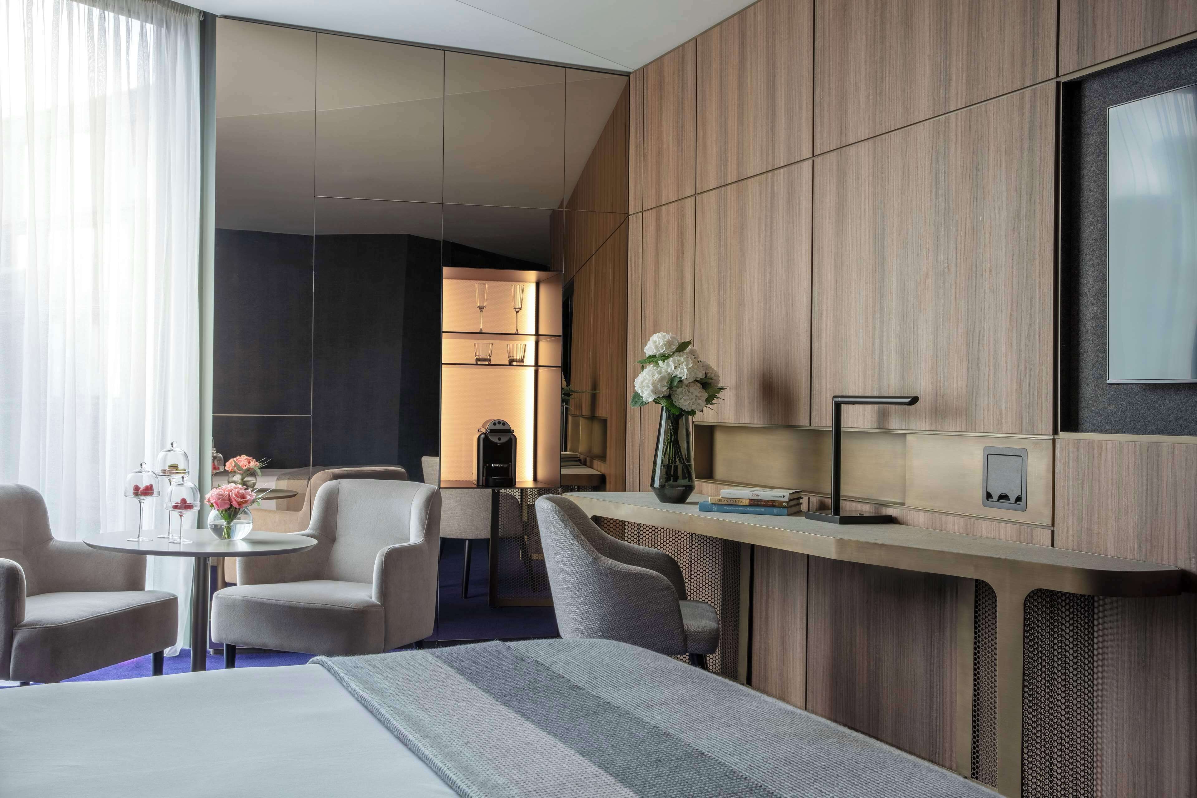 A modern hotel room with a large bed, a desk with a vase of flowers, a coffee machine, and a seating area with chairs.