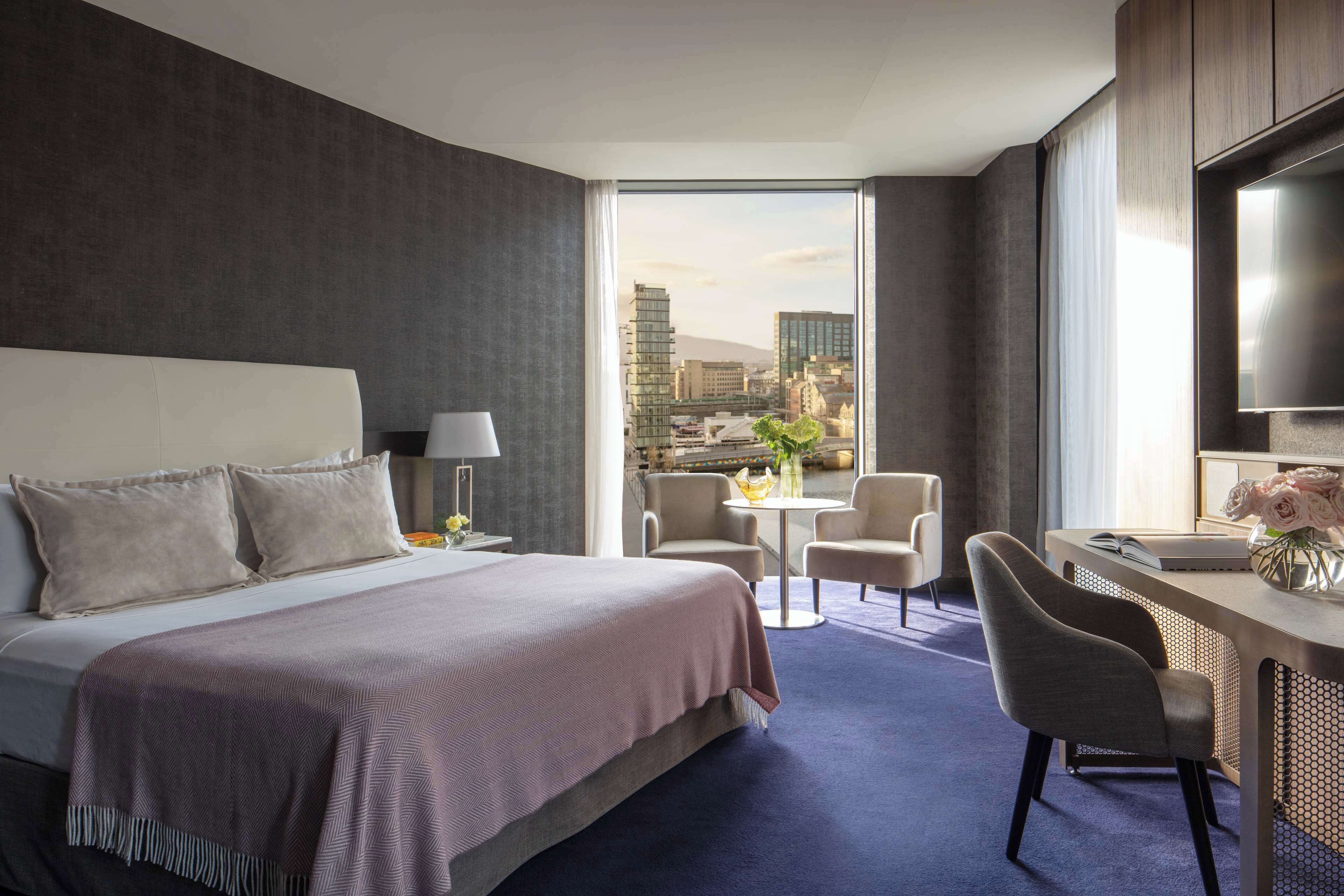 A modern hotel room with a large bed, a desk, and a view of a cityscape.