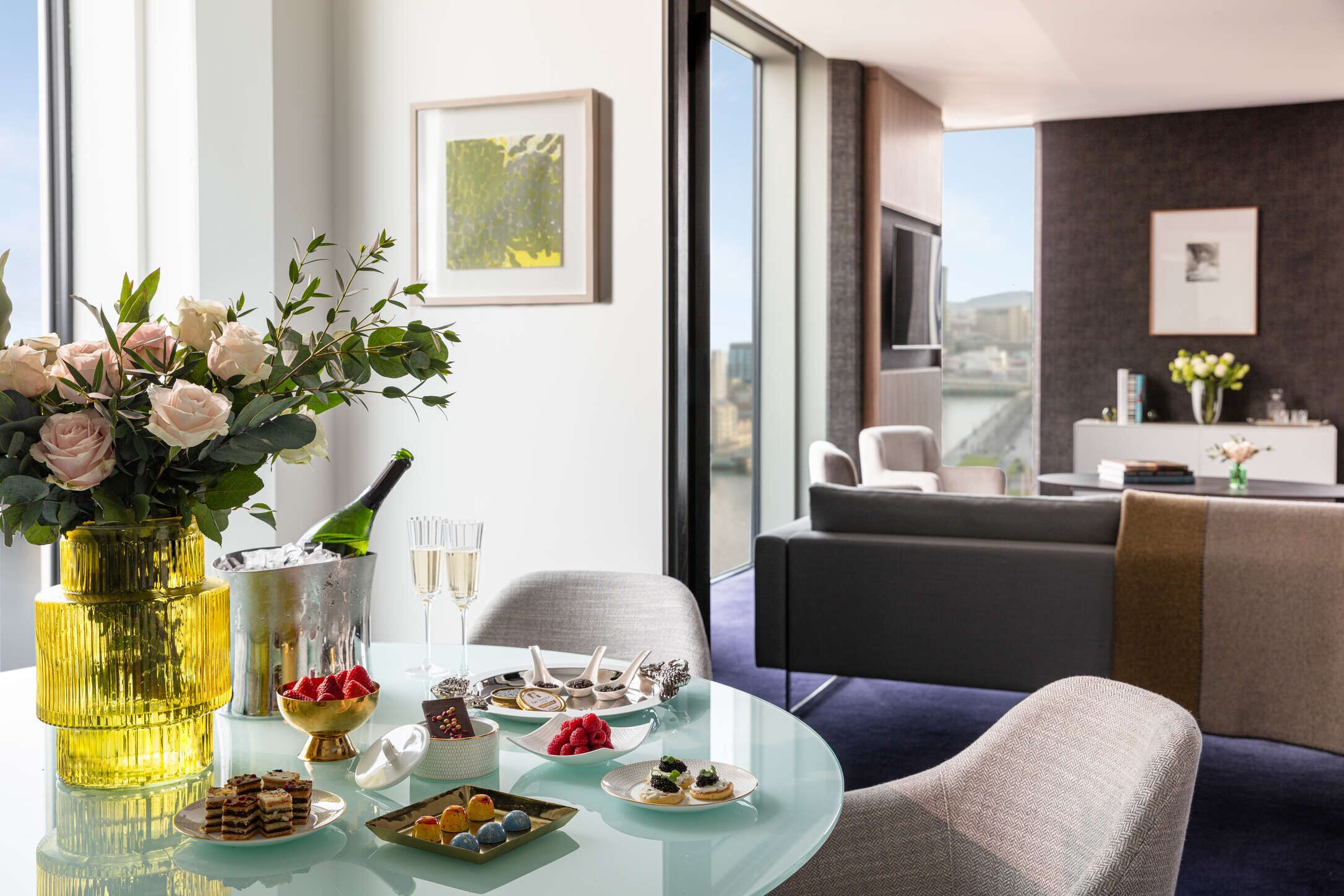 A modern dining area with a glass table, floral centerpiece, champagne, and various appetizers.