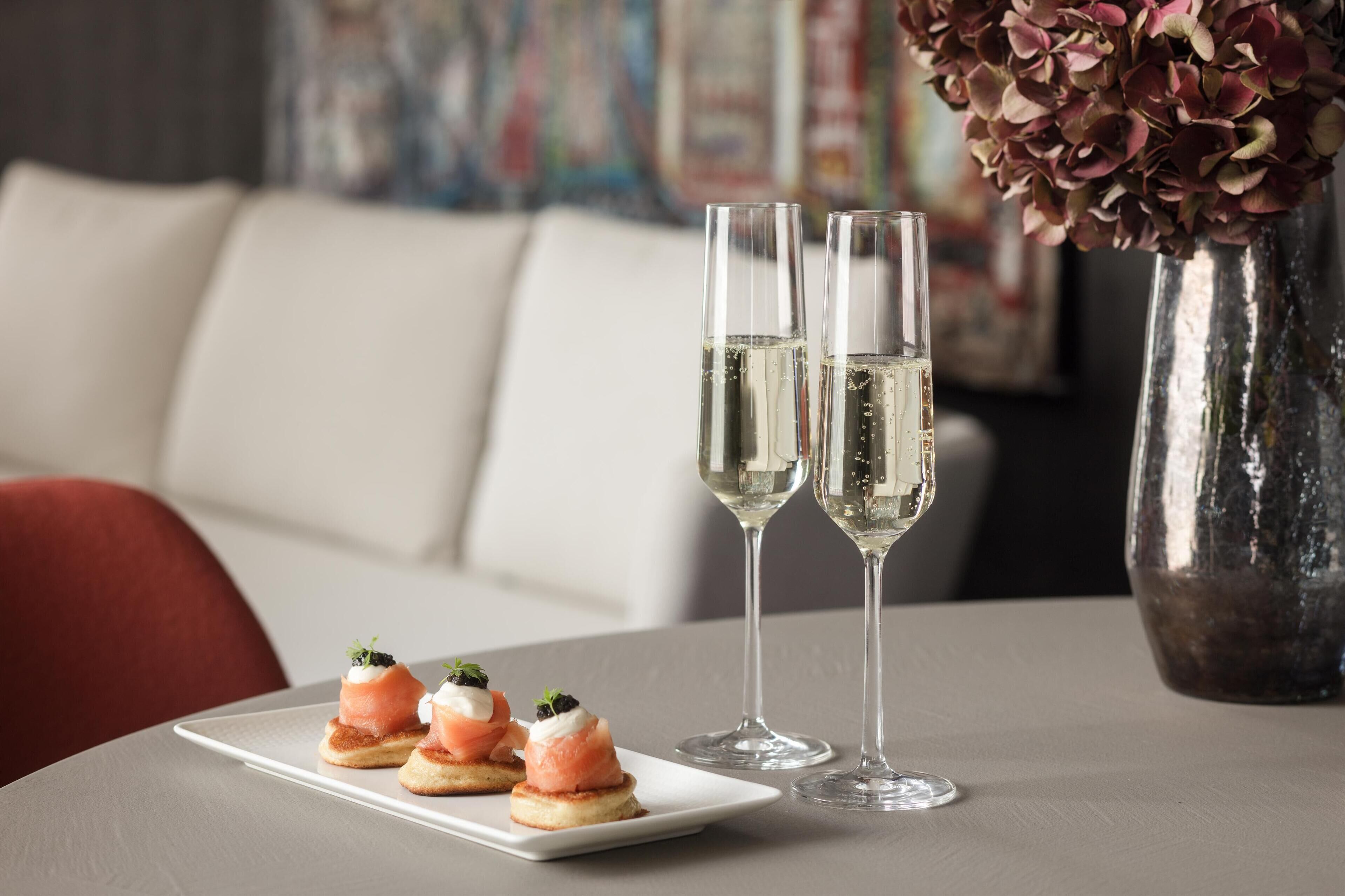 Une table avec deux verres de vin blanc, une assiette avec trois amuse-gueules et un vase rempli de fleurs.