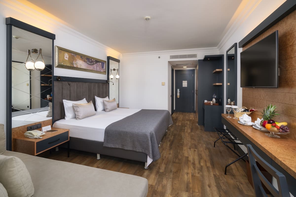 A modern hotel room with a large bed, bedside tables, a desk with a flat-screen TV, and a fruit bowl on the desk.