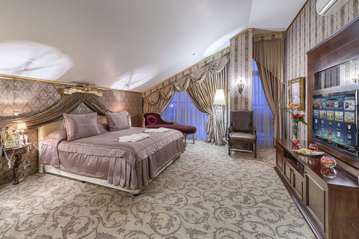 A luxurious bedroom with a large bed, a vanity with a mirror, a television, and a seating area with a chair and a table.
