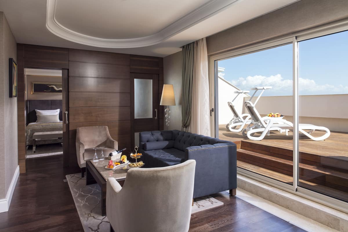 A modern hotel room with a large sliding glass door leading to a balcony. The room features a dark wood panel wall, a comfortable sofa, a dining table with a vase of flowers, and a bed with a view.