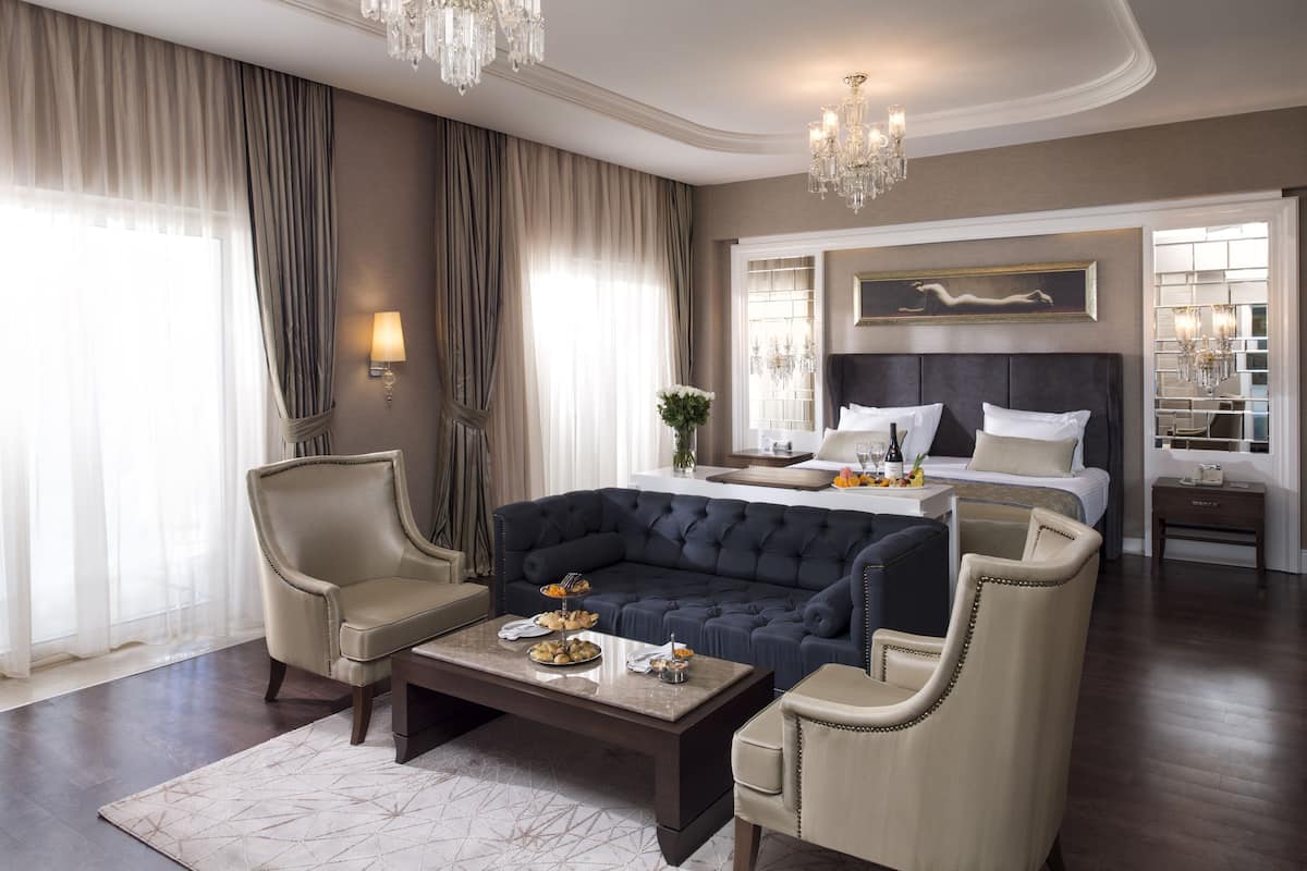 A spacious bedroom with a large bed, a dark blue sofa, two armchairs, a coffee table, and a chandelier.