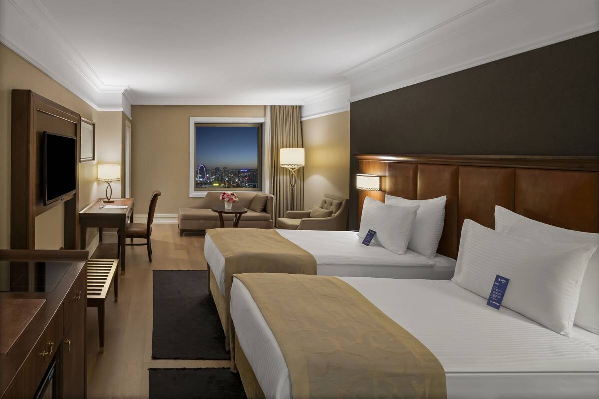 A hotel room with two beds, a desk, a TV, a window with a city view, and a chair.