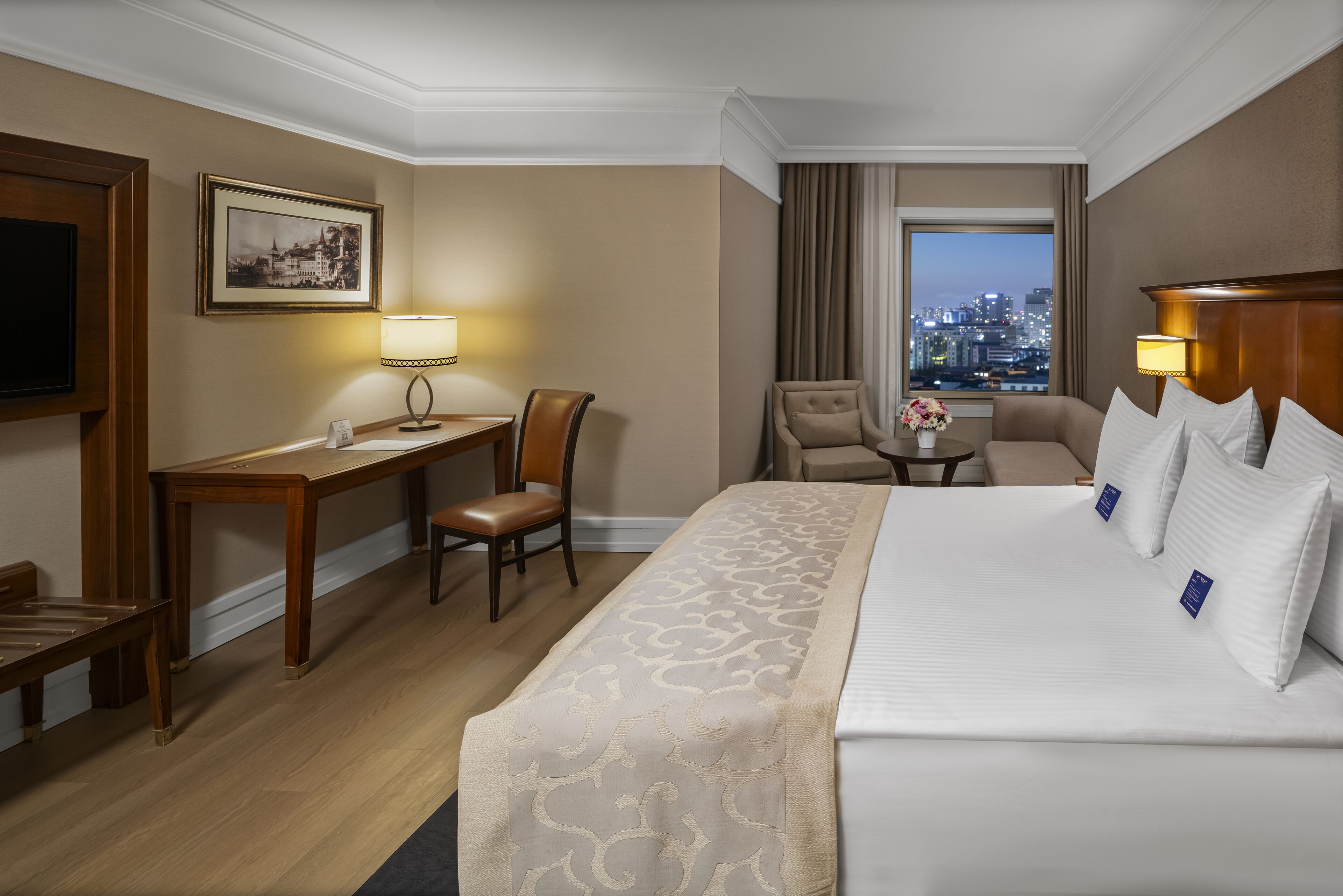 Habitación de hotel con una cama grande, un escritorio, una silla y vistas de la ciudad al atardecer.