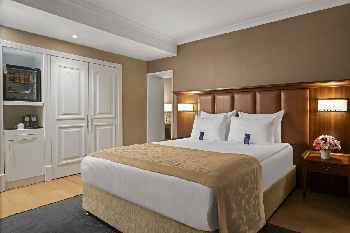 A hotel room with a large bed, a wooden headboard, bedside lamps, a nightstand with a vase of flowers, and a minibar.
