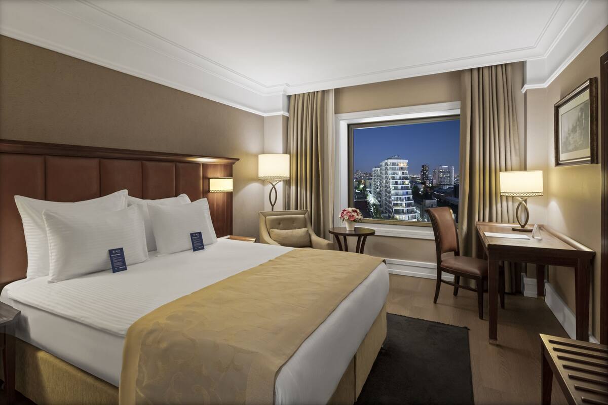 A hotel room with a large bed, two chairs, a desk, and a window with a city view.