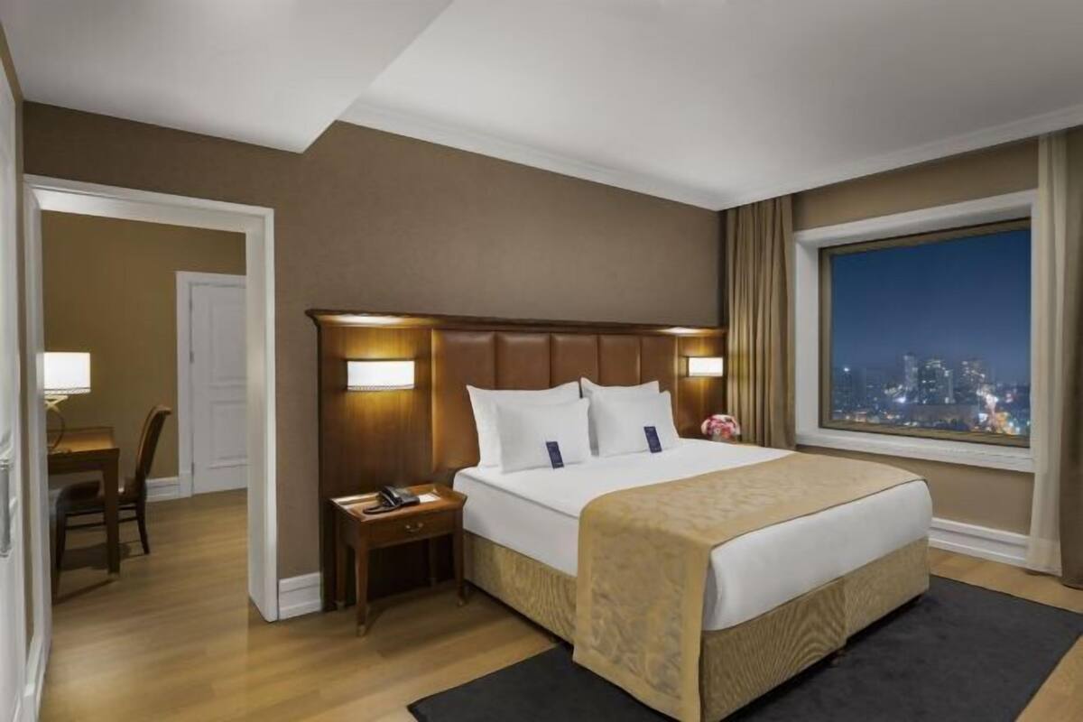A hotel room with a large bed, a nightstand, a desk, and a window with a city view.
