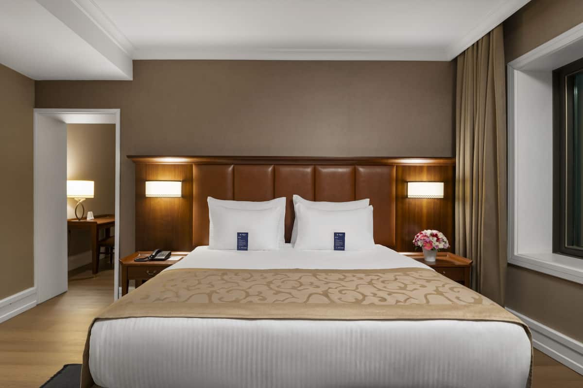 A hotel room with a large bed, two bedside tables with lamps, a wooden headboard, and a window with curtains.