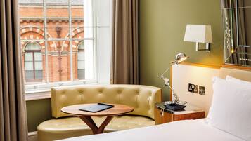 A hotel room with a bed, a small round table with a book, a nightstand with a lamp, and a view of a brick building through a window with curtains.