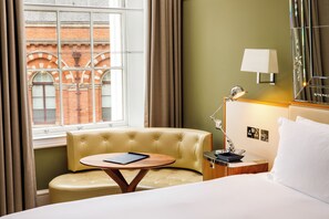 A hotel room with a bed, a small round table with a book, a nightstand with a lamp, and a view of a brick building through a window with curtains. - Kaya Great Northern Hotel (London)