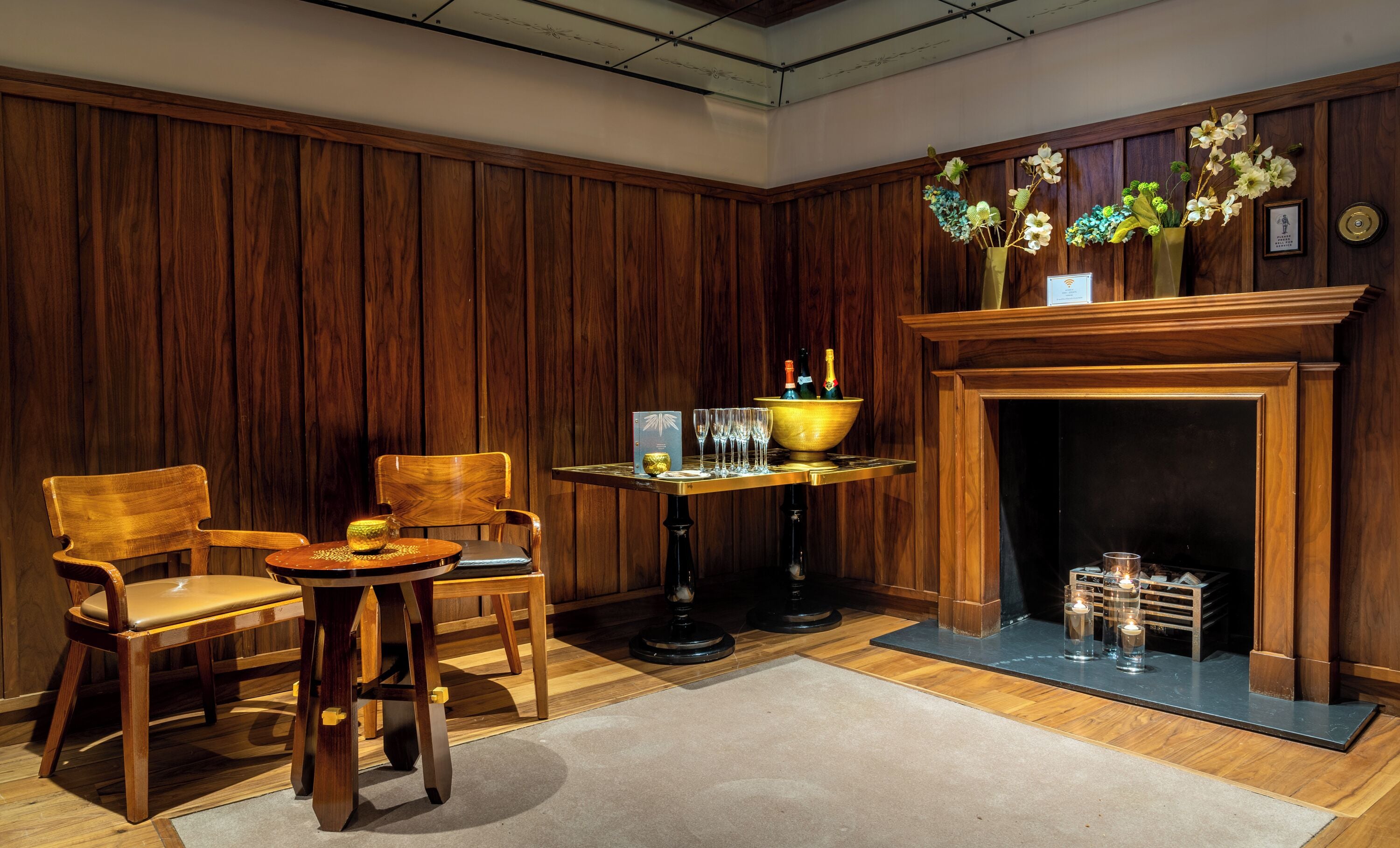 A room with wooden paneling, a fireplace, a table with glasses and bottles, and two chairs.