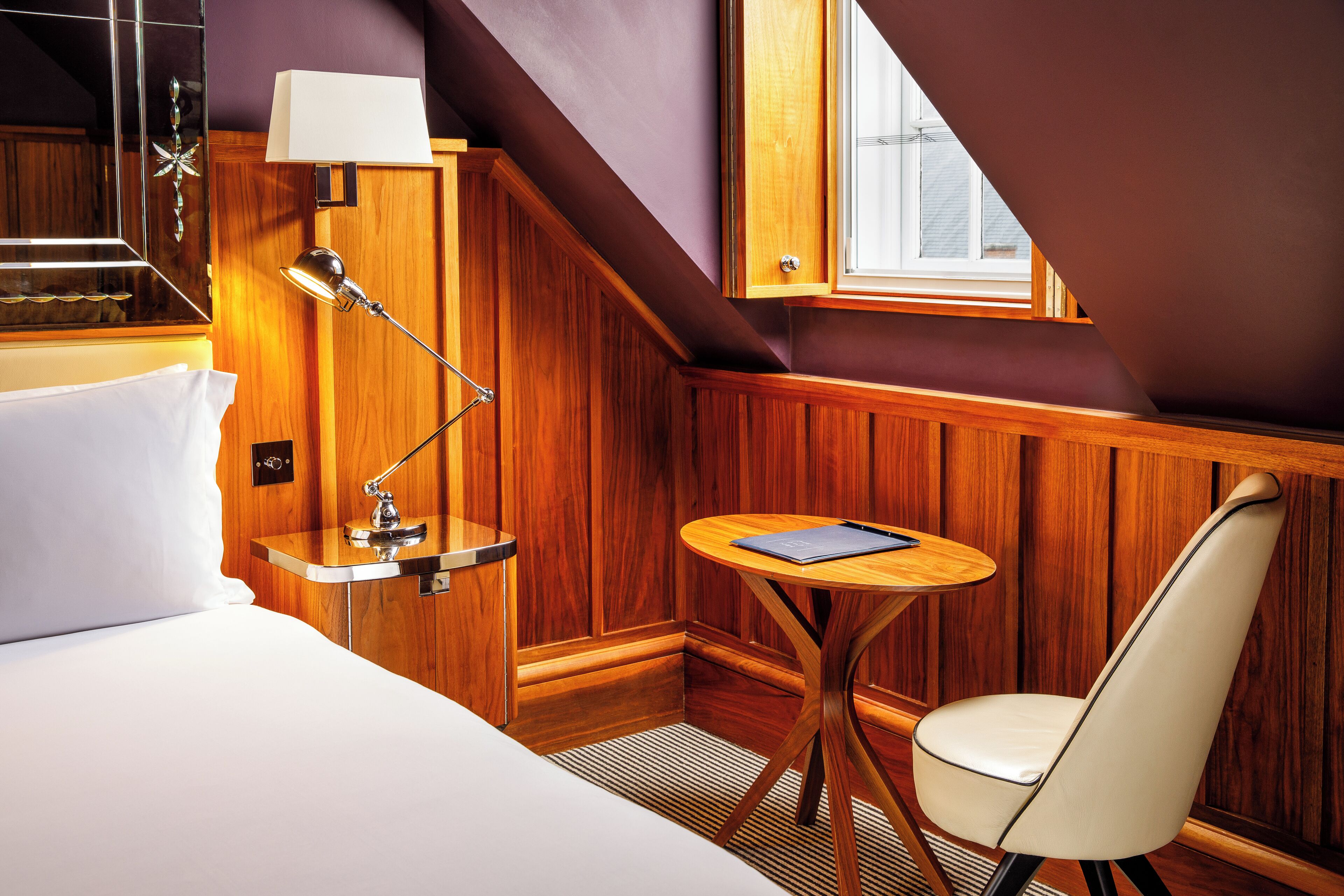 A hotel room with a bed, a bedside lamp, a small round table, and a chair.