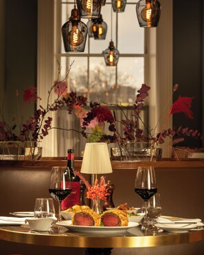 A dining table set with wine glasses, a bottle of red wine, a lamp, and a plate of steak with a decorative roll. - Kaya Great Northern Hotel (London)