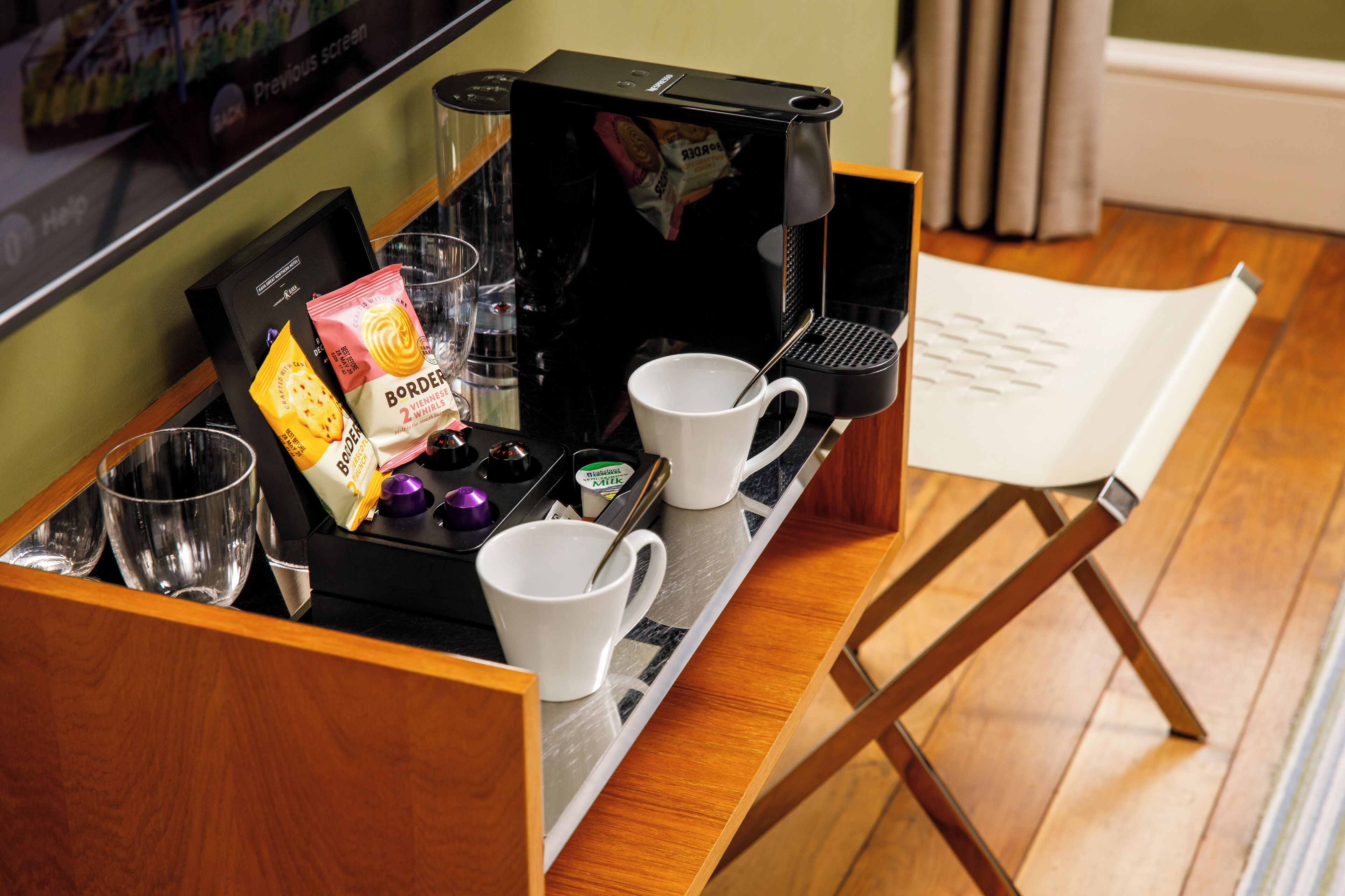 A coffee station with a coffee maker, two white cups, a glass, a packet of snacks, and a menu.